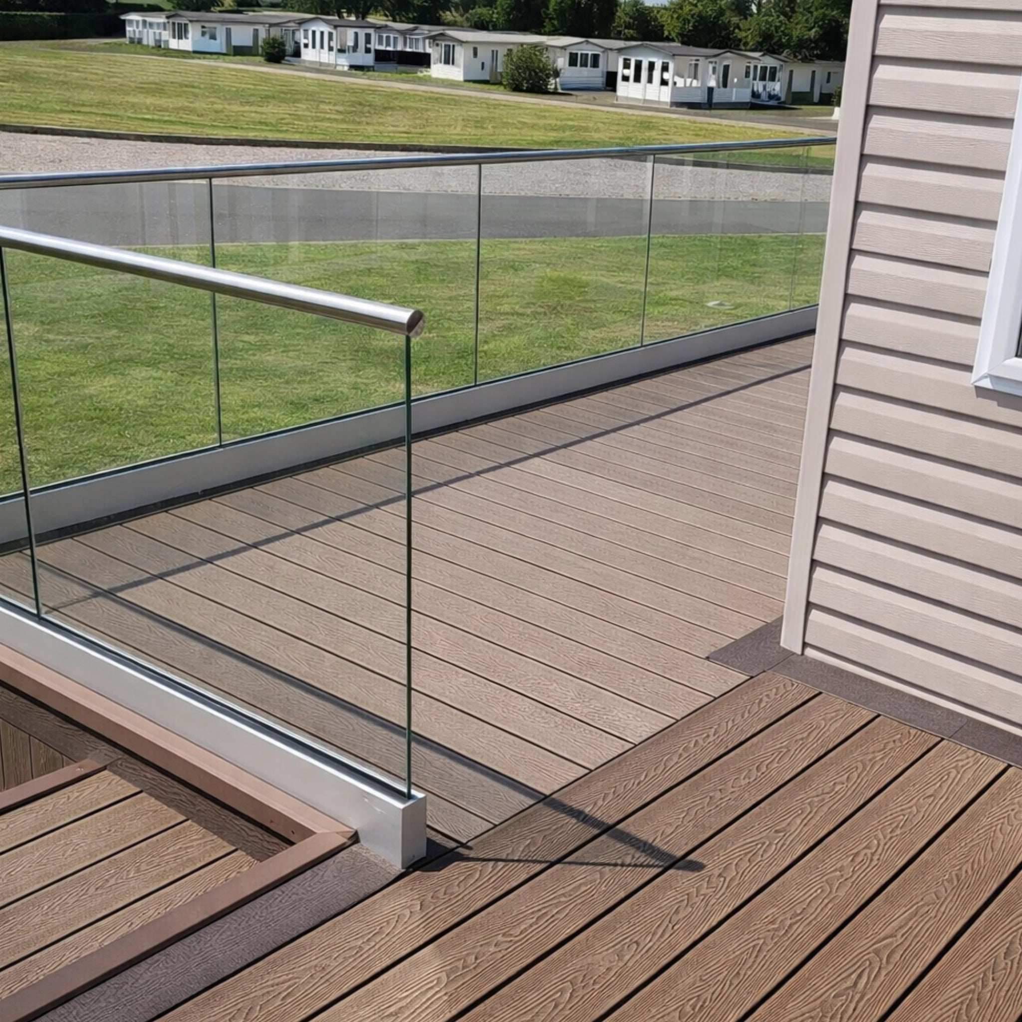 Wooden deck with glass railing overlooking a grassy area with mobile homes.
